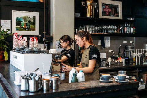 Two baristas working behind a counter in a coffee shop with various coffee-making equipment and decor.